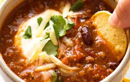 Corn chip being dipped into a bowl of Chili topped with sour cream, grated cheese and cilantro