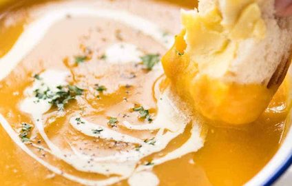 Close up of dunking crusty bread into thick and creamy pumpkin soup in a rustic white enamel bowl.